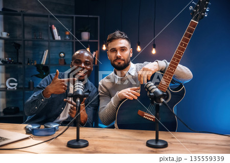 Posing, holding acoustic guitar. Two radio presenters recording an audio broadcast in a studio Posing, holding acoustic guitar. Two radio presenters recording an audio broadcast in a studio 135559339