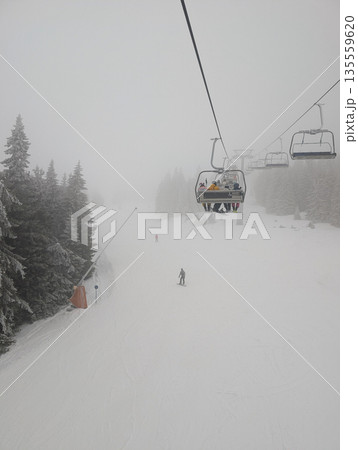 Chairlift carrying skiers above foggy slope in winter forest. Low visibility travel, alpine transport systems, and mountain recreation through elevation, motion, and seasonal conditions. 135559620