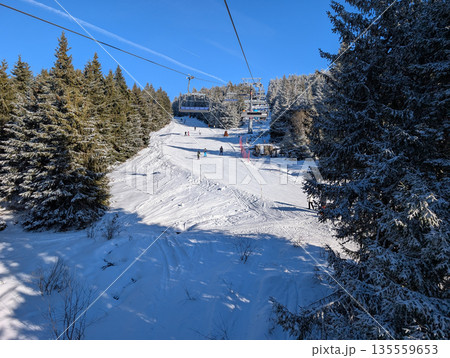Ski lift line above forested slope with riders in Kopaonik resort. Winter mobility, alpine tourism, and shared outdoor experience through snowy terrain and seasonal recreation. Ski lift line above forested slope with riders in Kopaonik resort. Winter mobility, alpine tourism, and shared outdoor experience through snowy terrain and seasonal recreation. 135559653