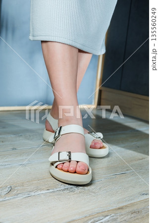 White leather sandals on women's feet. Female feet in comfortable casual sandals White leather sandals on women's feet. Female feet in comfortable casual sandals 135560249
