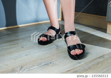 Black leather sandals on women's feet. Female feet in comfortable casual sandals Black leather sandals on women's feet. Female feet in comfortable casual sandals 135560313