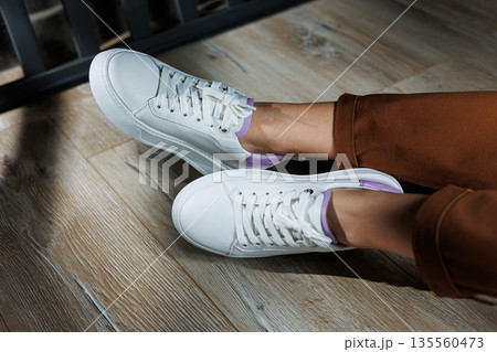 Comfortable female legs in white sneakers with flat soles. Close-up of female legs in leather sneakers. Women's summer sneakers. Collection of women's leather shoes Comfortable female legs in white sneakers with flat soles. Close-up of female legs in leather sneakers. Women's summer sneakers. Collection of women's leather shoes 135560473