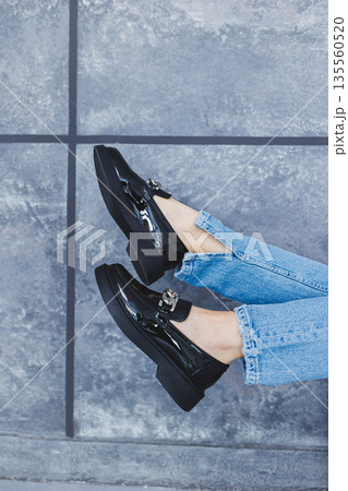 A young woman with slender beautiful legs in fashionable black leather shoes sits on a chair in the studio. Women's stylish loafers, close-up. Spring shoe collection 135560520