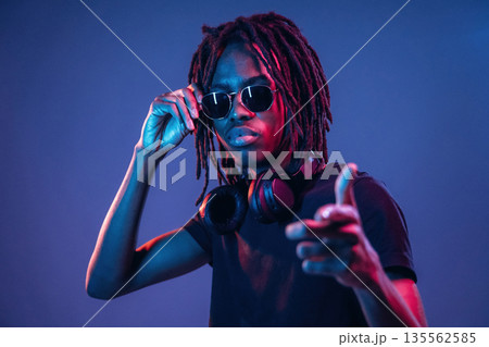 Pointing by finger, in sunglasses. African American guy is studio with neon lighting Pointing by finger, in sunglasses. African American guy is studio with neon lighting 135562585