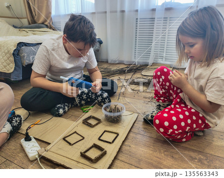 Two young children sit on the floor, intently working on a DIY project using cut tree branches, cardboard, and a glue gun. The children are developing their creativity and fine motor skills. Two young children sit on the floor, intently working on a DIY project using cut tree branches, cardboard, and a glue gun. The children are developing their creativity and fine motor skills. 135563343