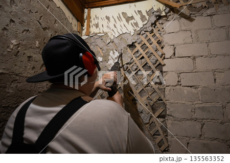 A construction worker performs demolition work using a hammer drill. A man uses a hammer drill to remove old plaster and paneling from an interior wall during a home renovation. A construction worker performs demolition work using a hammer drill. A man uses a hammer drill to remove old plaster and paneling from an interior wall during a home renovation. 135563352