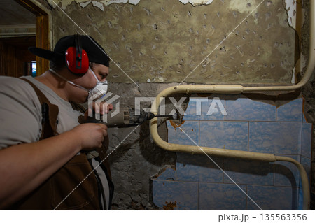 Construction worker wearing a respirator mask and earmuffs, using a hammer drill to remove old plaster and tiles from a dilapidated wall during a home renovation project. Construction worker wearing a respirator mask and earmuffs, using a hammer drill to remove old plaster and tiles from a dilapidated wall during a home renovation project. 135563356