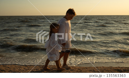 Children and brothers walking hand in hand along a sandy beach at sunset. The gentle lapping of the waves and the warm evening light evoke carefree summer memories of family togetherness and youth. 135563363