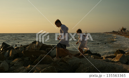 A cheerful brother and sister stroll and play on a rocky pier on a warm summer evening. The sunset over the calm ocean water illuminates everything around with a beautiful glow. 135563365