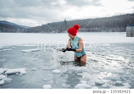 Senior woman breaking ice for winter swimming on frozen lake. 135563421
