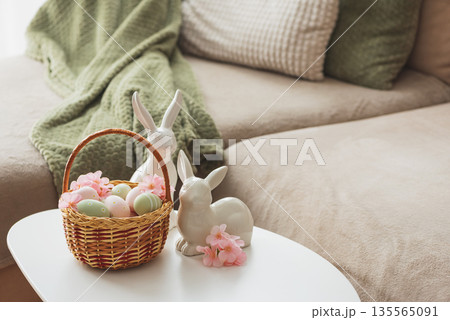 Easter Basket with Ester Eggs,  Plush Bunny and Spring Decor on White Table intoCozy Living Room 135565091