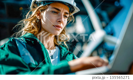 Female engineer wearing safety helmet and reflective jacket working on laptop at industrial facility or construction site 135565271