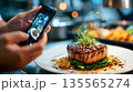 Hands using a smartphone to photograph a gourmet restaurant dish, highlighting modern food photography and social media culture 135565274