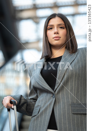 Young professional woman commuting or embarking on a business trip, standing with her luggage handle in what appears to be an airport or train station environment Young professional woman commuting or embarking on a business trip, standing with her luggage handle in what appears to be an airport or train station environment 135565423