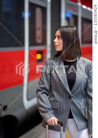 Young professional woman in a pinstripe blazer stands on a train platform with a suitcase, waiting confidently for a business trip in an urban commuter station Young professional woman in a pinstripe blazer stands on a train platform with a suitcase, waiting confidently for a business trip in an urban commuter station 135565424