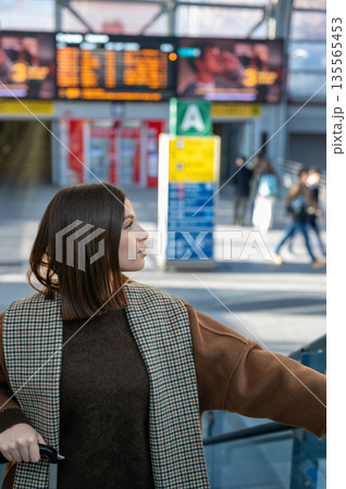 Young woman standing in bright airport terminal with luggage, looking toward departures board through large windows, poised and ready for an international journey or business trip 135565453