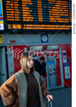 Woman standing in a train station, pulling a suitcase while checking the departures board, representing themes of travel, commuting, and urban transportation awaiting a journey 135565456