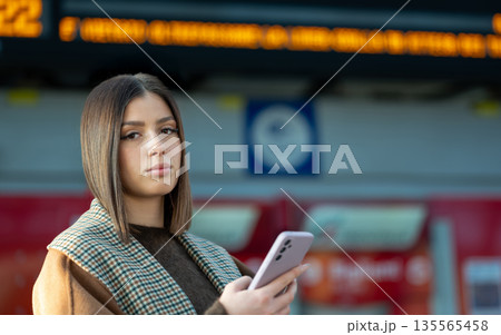 Young woman standing on a train station platform, holding her mobile phone and waiting for public transport, with a blurred information board in the background 135565458