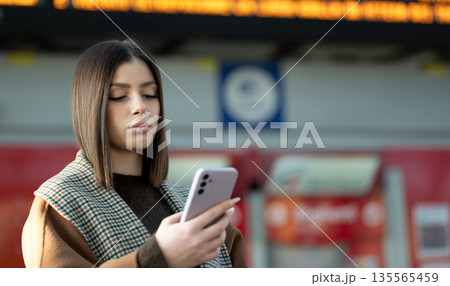 Young woman with brown hair in a coat checking her smartphone at a busy train station, waiting by the departure board and staying connected while commuting or traveling 135565459
