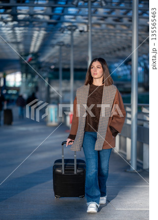 Young woman confidently walking through a modern airport terminal, pulling her black suitcase, representing travel, business trips, and vacation departures Young woman confidently walking through a modern airport terminal, pulling her black suitcase, representing travel, business trips, and vacation departures 135565463