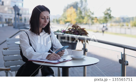 A business woman sits at a small table outside a cafe, focused on her phone and notes. She enjoys coffee while surrounded by a pleasant outdoor setting and warm sunlight. 135565751