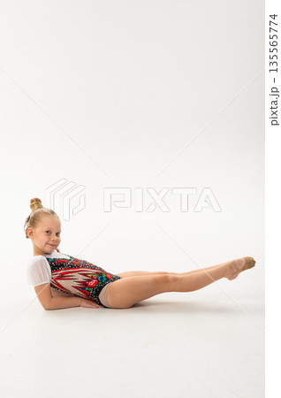 Smiling gymnast with cochlear implant stretching legs in studio leotard. Hearing support, inclusive physical therapy and childhood artistic development through controlled movement Smiling gymnast with cochlear implant stretching legs in studio leotard. Hearing support, inclusive physical therapy and childhood artistic development through controlled movement 135565774