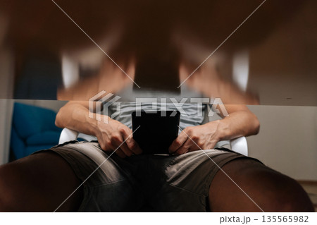 Unrecognizable man at home relaxing and engaging with electronic reader, immersing in digital content and enjoying quiet time, with blurred reflection showing hands and reader on table above. Unrecognizable man at home relaxing and engaging with electronic reader, immersing in digital content and enjoying quiet time, with blurred reflection showing hands and reader on table above. 135565982