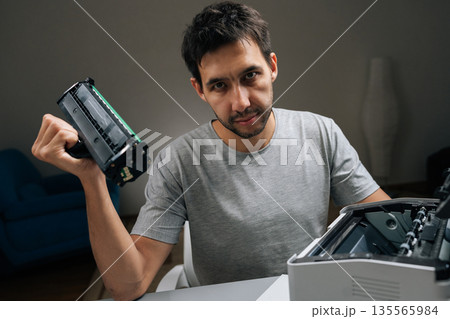 Young man holding empty toner cartridge from laser printer, looking directly at camera with frustrated expression while attempting to fix office equipment. Young man holding empty toner cartridge from laser printer, looking directly at camera with frustrated expression while attempting to fix office equipment. 135565984