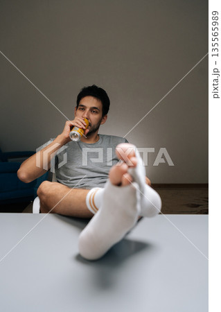 Full length vertical portrait of young man relaxing at home, having cold drink, feet with torn socks resting on table, representing daily life, comfort, leisure time, vulnerability after long day. Full length vertical portrait of young man relaxing at home, having cold drink, feet with torn socks resting on table, representing daily life, comfort, leisure time, vulnerability after long day. 135565989