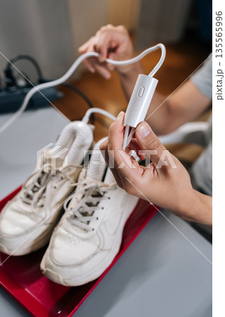 Vertical cropped shot of person plugging in portable electric shoe dryer to clean and dry white sneakers. Concept of footwear hygiene, maintenance, modern device technology for personal apparel care. 135565996