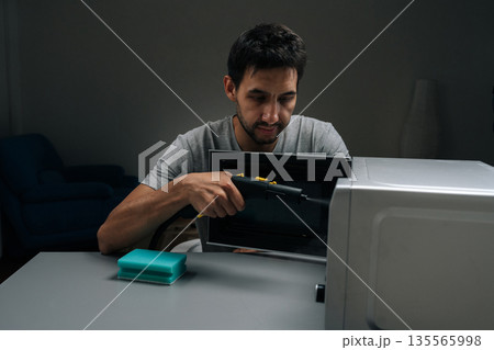 Man cleaning microwave interior with handheld steam cleaner, blasting hot vapor to lift grease and stains for fast, hygienic kitchen maintenance and sparkling results. Concept of kitchen hygiene. 135565998