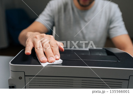 Close-up cropped shot of man performing printer maintenance, wiping dust off office laser printer, ensuring technology hygiene, cleaning and servicing electronic equipment in home office. 135566001
