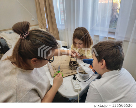 A mother and two children are engaged in creative activities, building a miniature house using a glue gun and natural materials. A family makes a craft for kindergarten or school. A mother and two children are engaged in creative activities, building a miniature house using a glue gun and natural materials. A family makes a craft for kindergarten or school. 135566520
