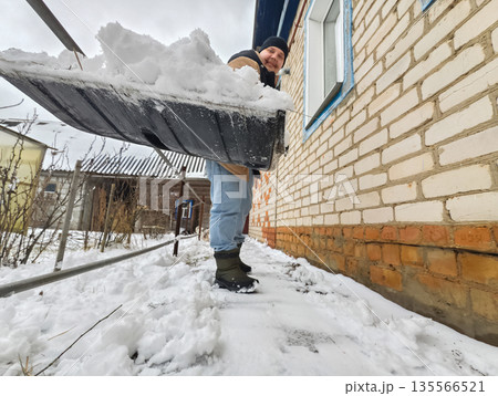 A man smiles as he shovels snow outside a brick house. A man is clearing snow from his yard in winter. Cheerful homeowner working outdoors. 135566521