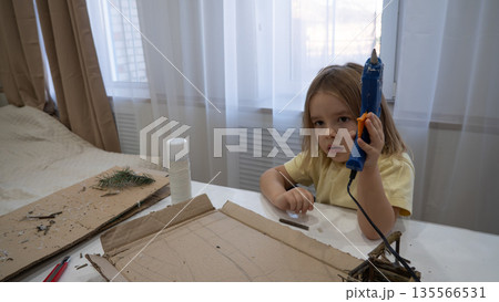 A child assembles a miniature cardboard house using natural materials and a glue gun at a bright kitchen table. The child focused on hands-on learning and creative play to create crafts. A child assembles a miniature cardboard house using natural materials and a glue gun at a bright kitchen table. The child focused on hands-on learning and creative play to create crafts. 135566531