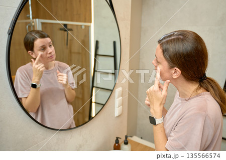 Woman standing in bathroom and touching face while looking at mirror Woman standing in bathroom and touching face while looking at mirror 135566574