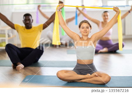 Group of multinational women and men sitting on sports mats and doing arm stretches using resistance bands at Pilates studio 135567216