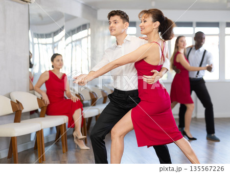 Hispanic guy and woman in red engrossed in passionate tango during dance class Hispanic guy and woman in red engrossed in passionate tango during dance class 135567226
