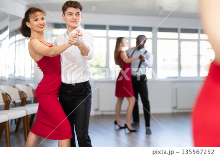 Positive woman in red dress and man practicing waltz dance moves in pair during group party in dancing studio Positive woman in red dress and man practicing waltz dance moves in pair during group party in dancing studio 135567252