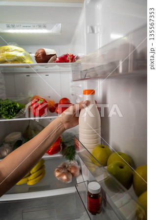 Hand reaching for a bottle of milk inside a refrigerator filled with fresh fruits and vegetables, showcasing a variety of colorful produce and organized shelves with food items Hand reaching for a bottle of milk inside a refrigerator filled with fresh fruits and vegetables, showcasing a variety of colorful produce and organized shelves with food items 135567353