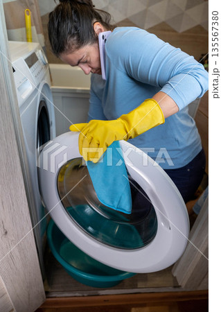 Woman wearing yellow gloves is cleaning the glass door of a washing machine while holding a smartphone in a modern laundry room with a blue bucket nearby 135567380