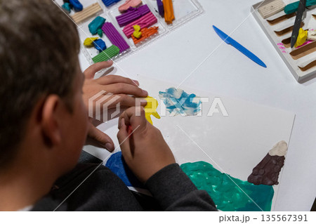 Child with short brown hair is painting a colorful landscape on white paper using yellow, blue, and green paint with modeling clay in the background during an art activity 135567391