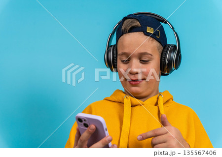 Young boy wearing headphones and a yellow hoodie is using a smartphone while smiling against a bright blue background with copy space for text or graphics Young boy wearing headphones and a yellow hoodie is using a smartphone while smiling against a bright blue background with copy space for text or graphics 135567406