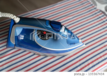 Electric steam iron in blue color resting on a striped ironing board with red and white lines, ready for use in a domestic laundry setting Electric steam iron in blue color resting on a striped ironing board with red and white lines, ready for use in a domestic laundry setting 135567434