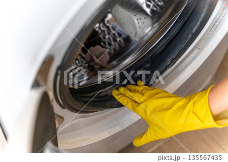 Close-up view of a hand wearing a yellow rubber glove inspecting the rubber seal of a washing machine door during routine maintenance in a home setting 135567435