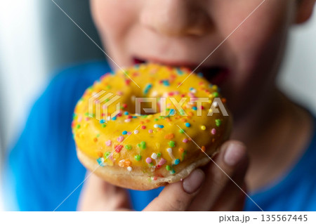 Child holding a yellow frosted donut with colorful sprinkles, preparing to take a bite, wearing a blue shirt, with a soft background creating a playful atmosphere Child holding a yellow frosted donut with colorful sprinkles, preparing to take a bite, wearing a blue shirt, with a soft background creating a playful atmosphere 135567445