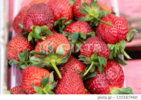 Fresh red strawberries with green leaves piled together in a clear container on a wooden surface, showcasing vibrant colors and natural textures for culinary use Fresh red strawberries with green leaves piled together in a clear container on a wooden surface, showcasing vibrant colors and natural textures for culinary use 135567460