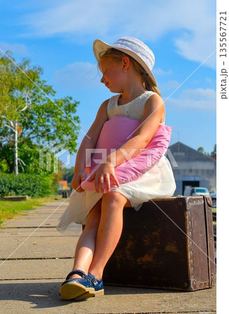 A cute little girl in a white dress and hat is waiting for her vacation. The little girl is waiting for the vacation train to arrive. The concept of a carefree childhood, summer vacation, and family. A cute little girl in a white dress and hat is waiting for her vacation. The little girl is waiting for the vacation train to arrive. The concept of a carefree childhood, summer vacation, and family. 135567721