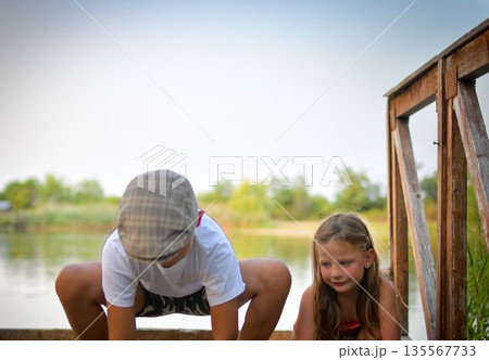 A little boy wearing a flat cap and a cute little girl are sitting on a wooden pier on the shore of a lake. Small children are sitting on a jetty at the edge of a pond. The concept of a carefree A little boy wearing a flat cap and a cute little girl are sitting on a wooden pier on the shore of a lake. Small children are sitting on a jetty at the edge of a pond. The concept of a carefree 135567733