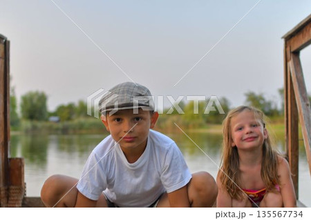 A little boy and cute girl kneel on a wooden pier on the shore of a lake. Small children sit on a pier at the edge of a pond. The concept of a carefree childhood and summer vacations. A little boy and cute girl kneel on a wooden pier on the shore of a lake. Small children sit on a pier at the edge of a pond. The concept of a carefree childhood and summer vacations. 135567734